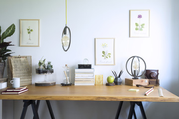Modern home office interior with wooden desk, books,poster illustrations of plants, table lamp and office accessories. Stylish creative and vintage desk.