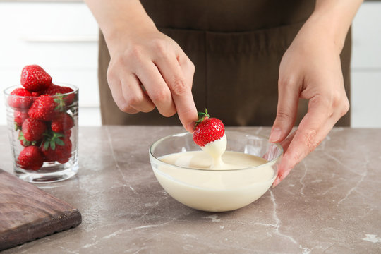 Woman dipping ripe strawberry into bowl with white melted chocolate on table