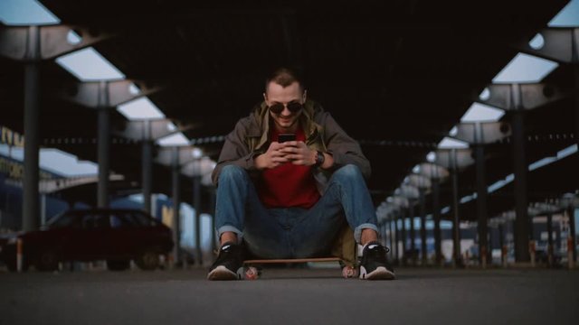 Attractive stylish guy in sportive clothes sitting on skateboard, looking at smartphon, browsing. Handsome Caucasian smiling young man in sunglasses texting. Outdoors.