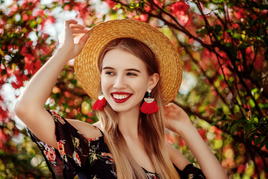 Young Beautiful Happy Smiling Girl With Red Lips, Long Blonde Hair, Wearing Straw Hat, Long Tassel Earrings, Cold Shoulder Dress, Posing Near Blooming Tree