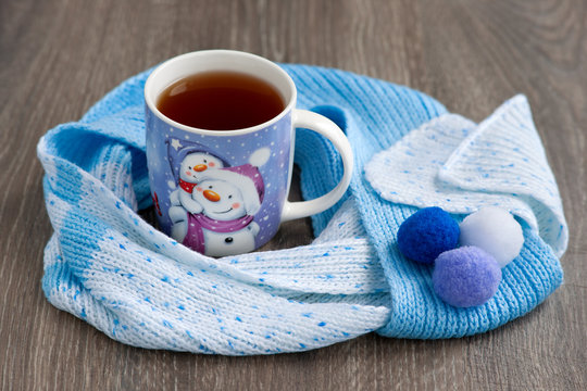 Mug of tea with a picture of a snowman for a cold winter. A hot tea mug is wrapped in a warm knitted scarf. Hot tea creates coziness in winter.