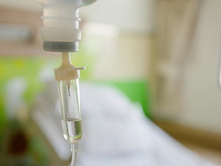 Closeup of saline drop in an infusion set's drip chamber infusing fluids, medication, and nutrients into a circulatory system of a patient, with a defocus of hospital patient room in the background