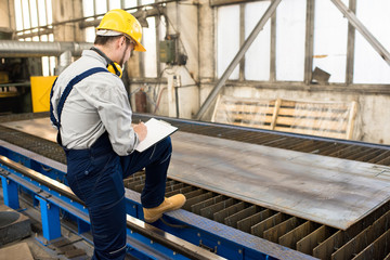 Naklejka na ściany i meble Serious concentrated factory inspector leaning by foot on conveyor belt and analyzing quality of metal sheer on rolling mill in shop while making notes