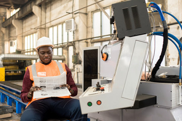 Serious relaxed handsome African-American machine operator in hardhat and vest sitting on chair and reading newspaper while automated machine working in plant shop