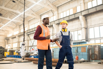Serious confident handsome African-American inspector in orange vest and hardhat using tablet while talking to construction worker and making report online
