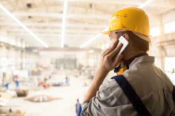 Rear view of serious busy construction engineer in yellow hardhat talking on mobile phone and...