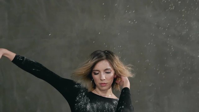 Slender caucasian blonde girl in black t-shirt and shorts is dancing in white powder cloud or dust indoors at gray wall background. Splashes flour or white powder on a young woman who dancing in the