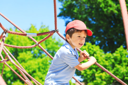 Cute and happy little four years old child boy climbed on top of the rope web on playground. Child crawling on rope mesh at park. A view of kid face with fancy red cup in play area.