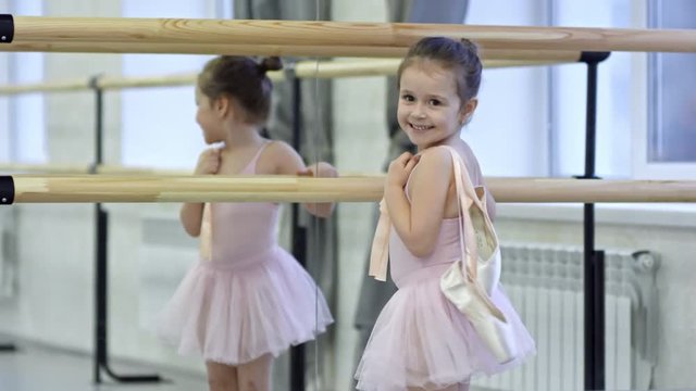 Medium shot of adorable little girl in pink leotard and tutu skirt carrying pointe shoes and running towards ballet barre in dance studio, then looking at camera and smiling