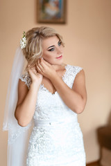 Fototapeta premium Beautiful bride in puts on earring. Beauty model girl is wearing jewelry for marriage. Wedding female portrait. Woman with curly hair and lace veil. Cute lady indoors, selective focus