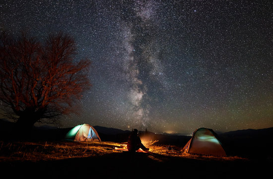 Night camping in mountains. Silhouette of male hiker sitting between two illuminated tents, enjoying campfire, beautiful starry sky and Milky way on background. Tourism, outdoor activity concept