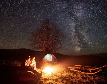 Camping night in mountains. Couple hikers boy and girl resting near burning campfire under starry sky with Milky way. Glowing tent, big tree and distant hills on background. Tourism and travel concept