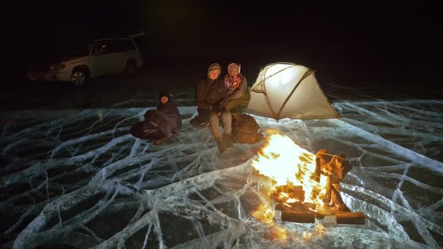 Four travelers by fire right on ice at night. Campground on ice. Tent stands next to fire. Lake Baikal. Nearby there is car. People are warming around campfire and are dressed in sleeping bags
