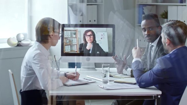 View through glass wall of multi-ethnic team of business professionals sitting in the office and video calling on computer with female colleague at meeting