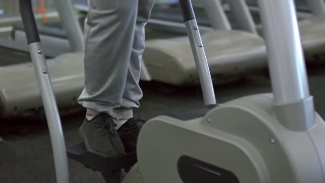 Tilt up of unrecognizable man in sweatpants and sneaker doing cardio on stepper machine in gym