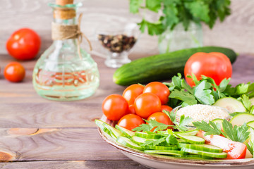 Fresh slices of cucumbers, tomatoes, sesame seeds in a bowl and parsley leaves on a plate on a wooden table. Oil and seasoning for cooking vegetable salad