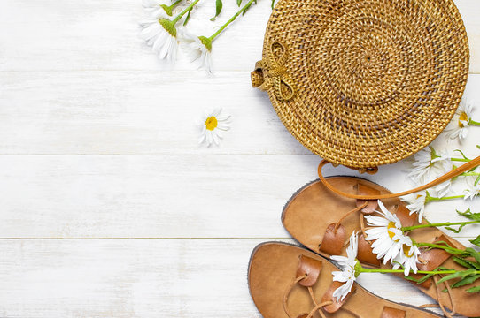 Fashionable handmade natural organic rattan bag, leather sandals, chamomile flowers on light wooden background. Copy space, top view. Ecobags from Bali. Eco-bag concept.