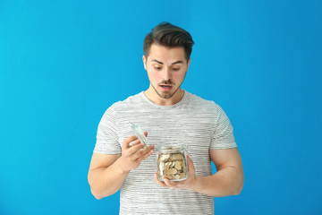 Young surprised man holding glass jar with coins on color background. Savings money concept