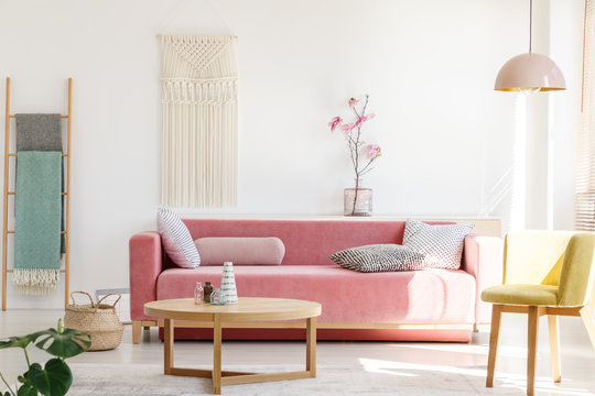 Yellow chair next to pink sofa and wooden table in pastel living room interior with flowers. Real photo