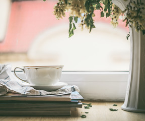 Cup on a stack of magazines at window with vase and flowers. Summer still life. Cozy home scene