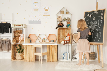 Little girl drawing on a blackboard in her room with wooden boxes, teddy bears and wall stickers © Photographee.eu