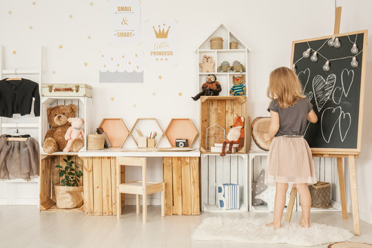 Little girl drawing on a blackboard in her room with wooden boxes, teddy bears and wall stickers