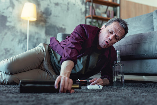 Bottle of vodka. Drunk alcohol addicted man reaching his hand after bottle of vodka while lying on the floor near cigarettes
