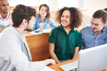 Obraz premium Cheerful excited multiethnic groupmates sitting at university desk ad discussing project while preparing report on laptop in classroom
