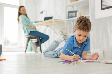 Painting on floor. Cute little smart boy feeling entertained while lying on the floor painting with...