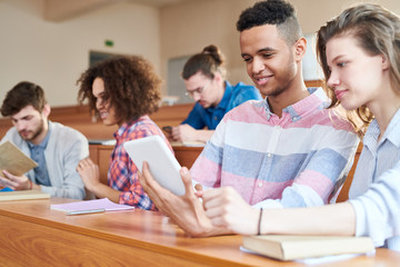 Fototapeta na wymiar Smiling positive young multiethnic groupmates in casual clothing sitting at university desk and watching educational video on tablet while learning subject together
