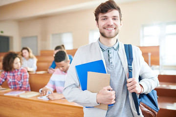 Cheerful positive ambitious handsome young guy in casual jacket smiling at camera and holding satchel and workbooks in lecture room - obrazy, fototapety, plakaty