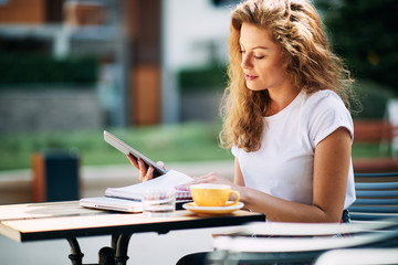 Female student using tablet and drinking coffee in cafe. Books and notebooks on a table.