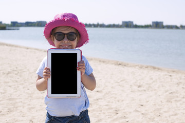 Little girl with a tablet computer in hands. Good summer weekend on the beach. Copy spce