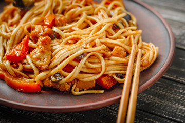Wok udon stir fry noodles with chicken and vegetables in plate and chopsticks on black wooden background