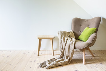 Beige armchair with a green pillow, blanket and a modern wooden table. Empty white wall in simple...