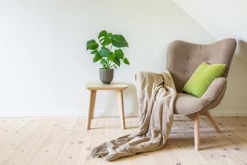 Beige armchair with a green pillow, blanket and a wooden table with a potted plant, fruit salad tree (Monstera deliciosa). Empty white wall in simple living room interior. Copy space © Forenius