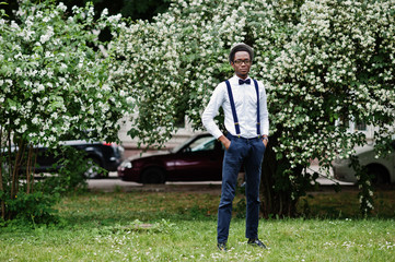 Stylish african american business man at pants with suspender and shirt with bow tie, hat and glasses posed outdoor.