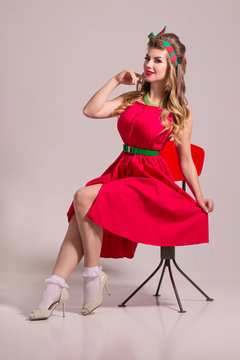 Smiling girl in red dress with hairdo sits on chair in grey studio, pin up style
