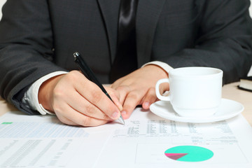 Hands of writing businessman sitting at table with cup, documents, diagrams