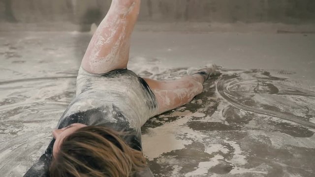 Dancer girl performs a modern dance lying on the floor in a cloud of powder or flour or dust. Young woman in a black body suit dancing contemporary dance on the floor in the studio