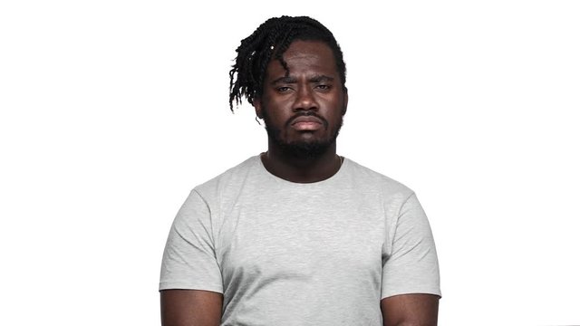 Portrait of angry dark skinned guy in basic outfit asking to keep quiet with strict gaze putting index finger on lips, isolated over white background slow motion. Concept of emotions