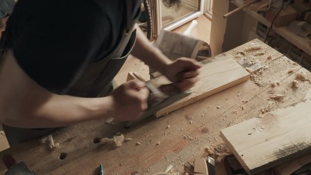 Skilled craftsman carpenter whittles workpiece of wooden board with jack-plane in a workshop. Making handmade furniture.