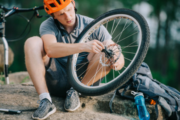 serious young trial biker fixing bicycle wheel outdoors while sitting on stone