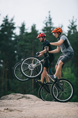 Fototapeta premium handsome young trial bikers standing on back wheels on rocky cliff with blurred pine forest on background