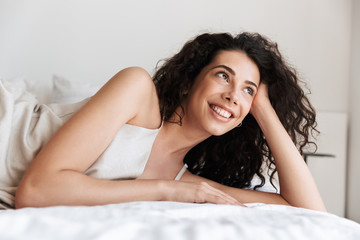 Naklejka premium Closeup photo of pretty young woman 20s with long curly hair wearing silk leisure clothing lying in hotel bed, and looking aside while propping up her head with hand