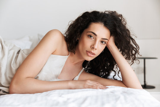 Closeup photo of beautiful young woman 20s with long curly hair wearing silk leisure clothing lying in bed at home, and looking at you while propping up her head with hand
