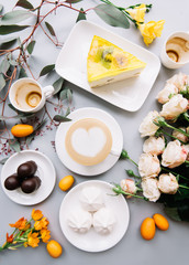 Breakfast flat lay: delicious fresh morning cappuccino coffee with a heart latte art on top, yellow cheese cake, marshmallows, chocolate, roses bouquet, eucalyptus and two empty cups, top view