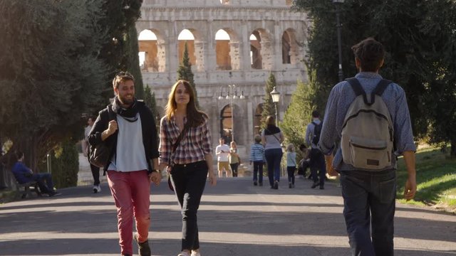 Young happy couple walk in park road and attractive young man glance to girlfriend gelousy with trees colosseum in background in rome at sunset lovely beautiful girl with long hair holding hands slow
