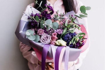 Very nice young brunette woman holding a big beautiful blossoming flower bouquet of roses, mattiola, carnations, eustoma, eucalyptus in purple colors on the grey wall background