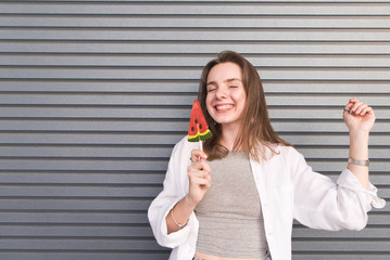 Happy girl standing with a lollipop in her hand and smiling warmly on a gray background. Portrait of smiling happy girl with candy on the background of the wall
